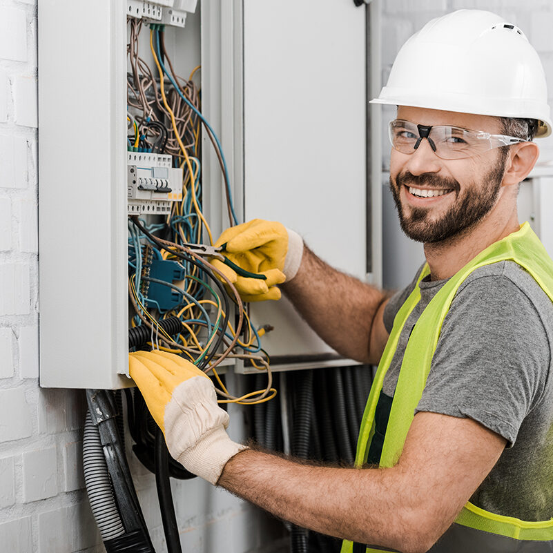 smiling handsome electrician repairing electrical box with pliers in corridor and looking at camera